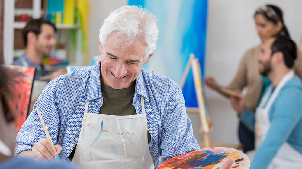 Senior man in group art class smiles at his painting while wearing frock and carrying painting tools