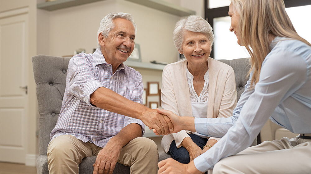 Senior living staff member shakes hand with smiling senior man seated next to his wife