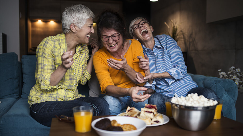 Three friends break out in laughter on couch together in dark theater room in front of plate of appetizers
