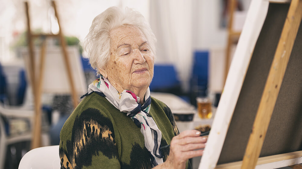 Elderly woman in green sweater with scarf sketches at canvas on easel in art room