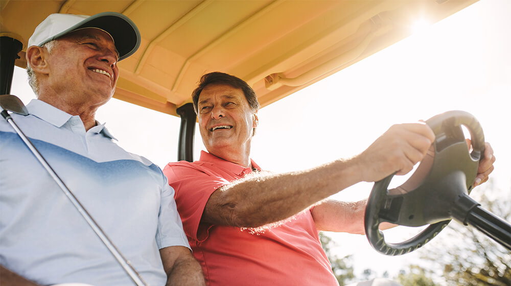 Two older men drive golf cart and smile at shared joke on sunny golf course