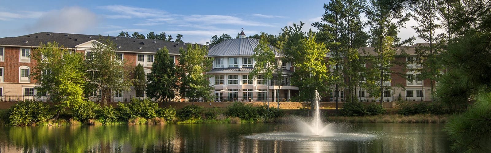Rear of the Georgian Lakeside building, surrounded by majestic trees and a lake with a fountain shooting up.
