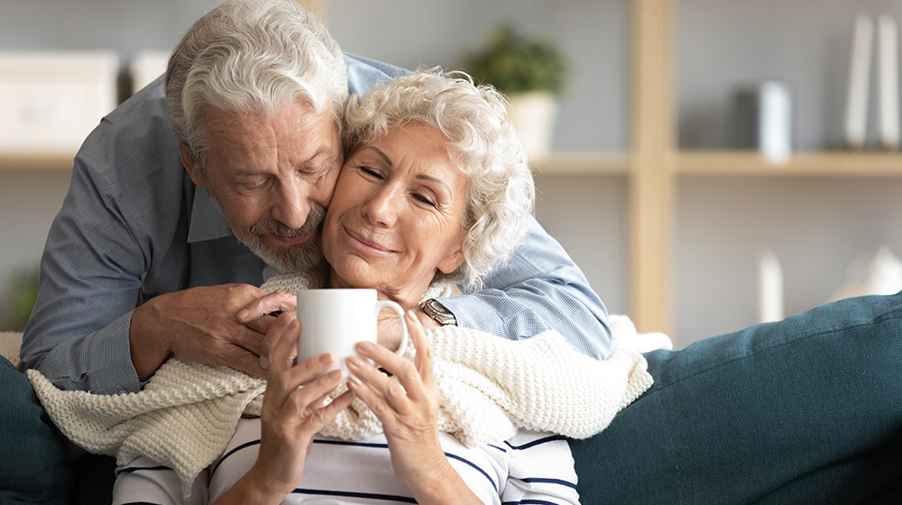 Caring elderly husband hugs wife brings tea